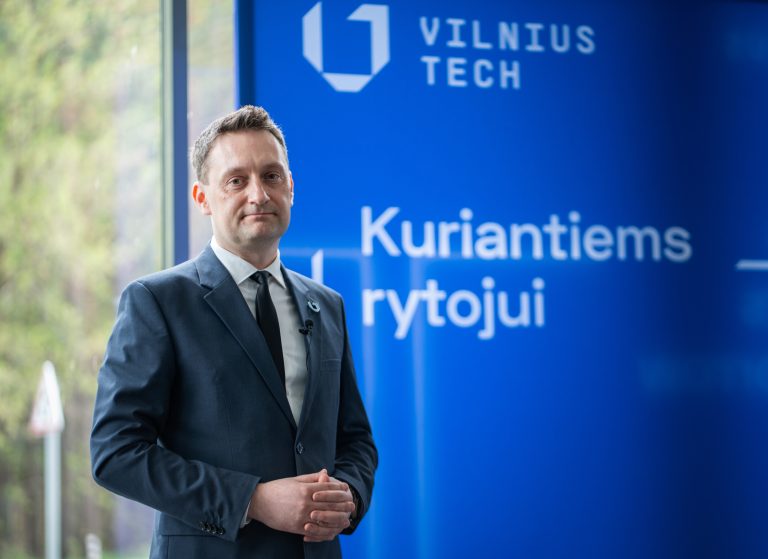 A man in a dark suit and black tie stands in front of a blue backdrop. He is posing formally, representing the SustainLivWork project.