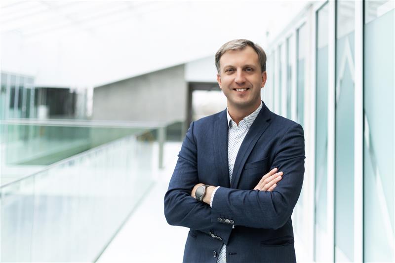 Color portrait of a man in a navy blazer and patterned button-up shirt, smiling at the camera with arms crossed, featured as part of the SustAInLivWork Centre of Excellence team advancing AI for sustainable living and working.
