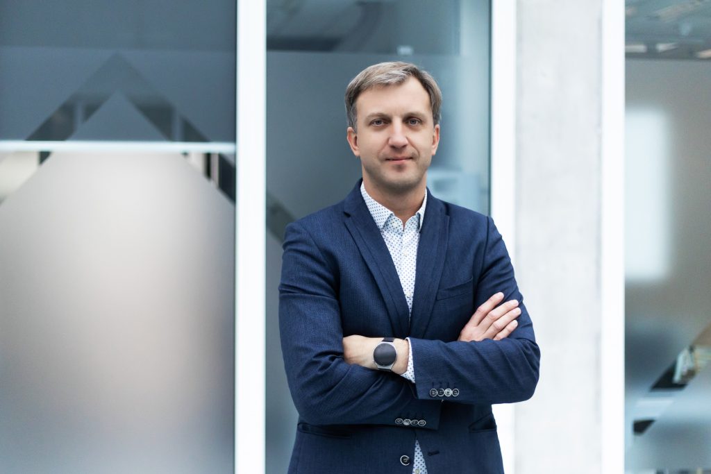 Color portrait of a man in a navy blazer and patterned button-up shirt, looking at the camera with a neutral expression and arms crossed, featured as part of the SustAInLivWork Centre of Excellence team advancing AI for sustainable living and working.