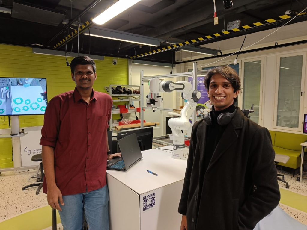 Two men stand in a robotics lab beside a white robotic arm mounted on a workstation with a laptop and QR code sticker visible on the table. The lab features bright overhead lights, green accent walls, computer monitors, and technical equipment, highlighting a collaborative engineering or research environment.
